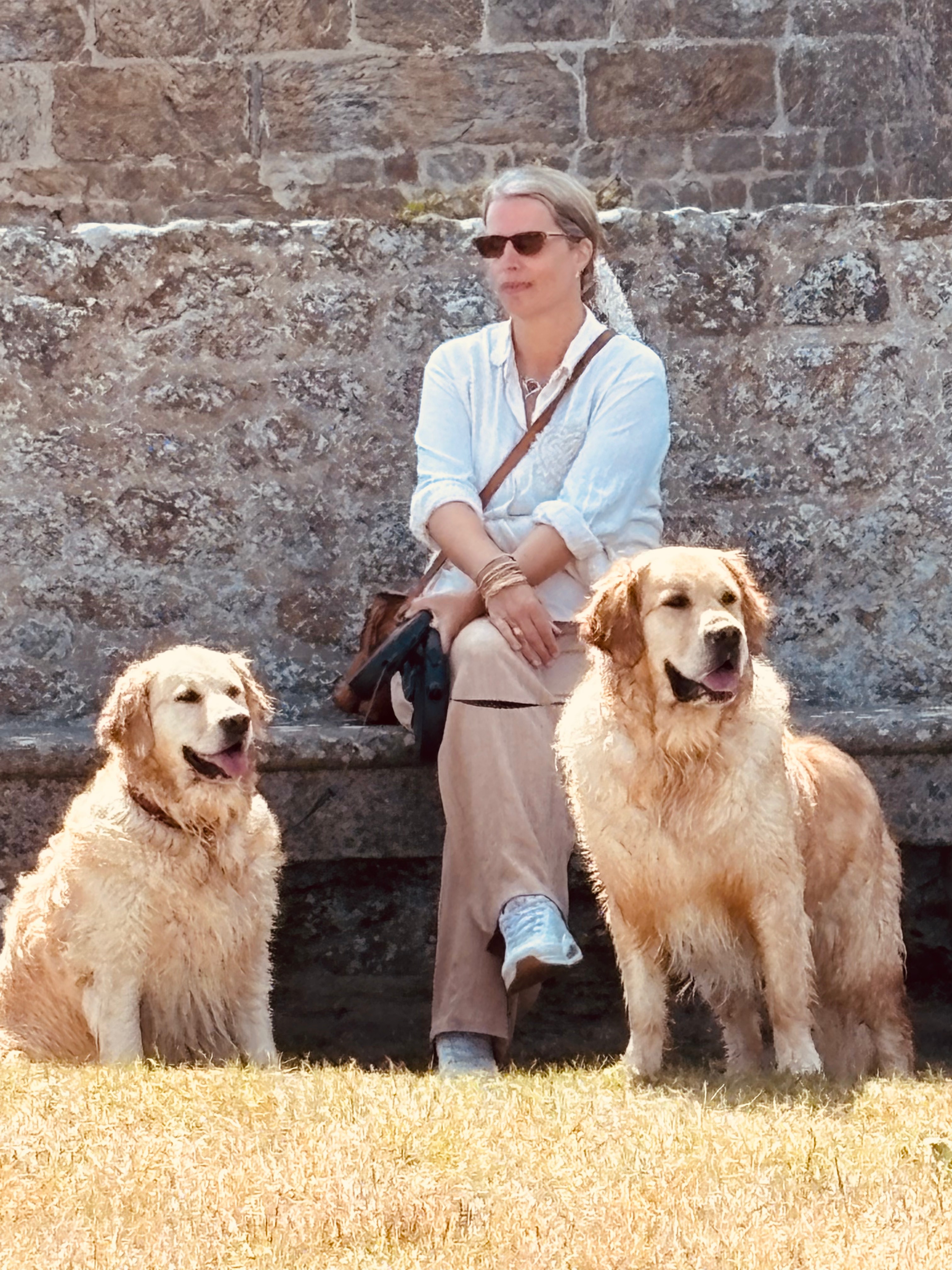 Een vrouw zit op een bank, gekleed in een witte blouse en met zonnebril, met aan weerszijden twee natte golden retrievers op een grasveld.